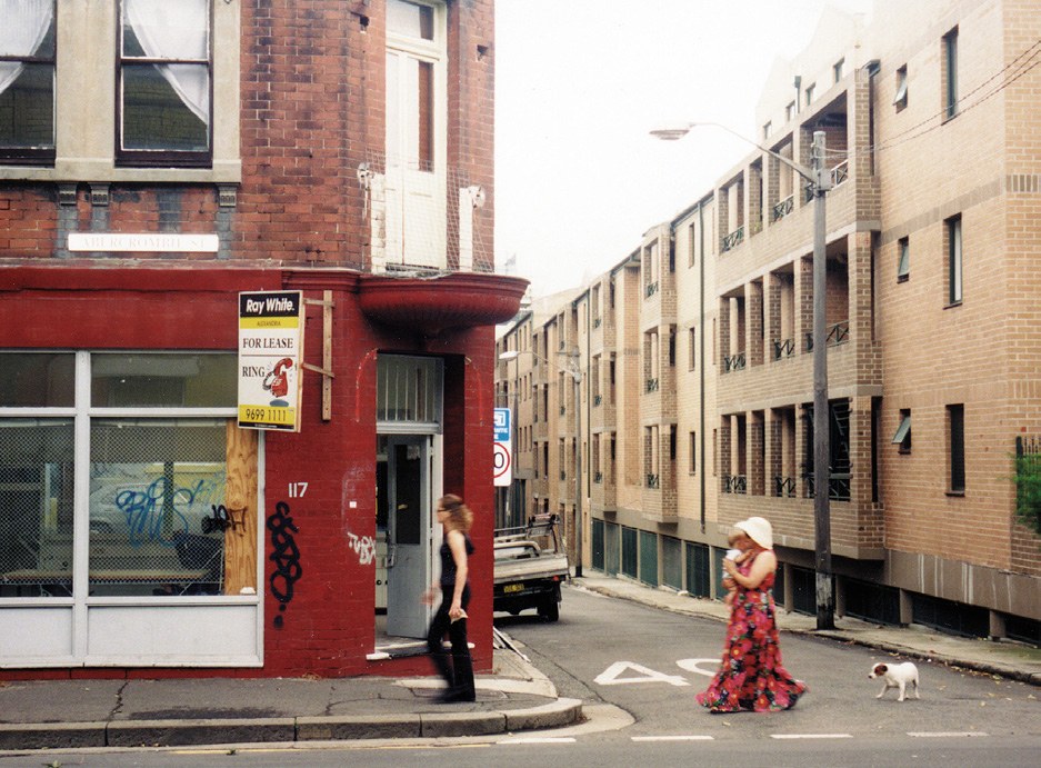The corner shopfront at 117 Abercrombie Street, Chippendale, in 2000, with a woman in a patterned dress and a small dog crossing the road.